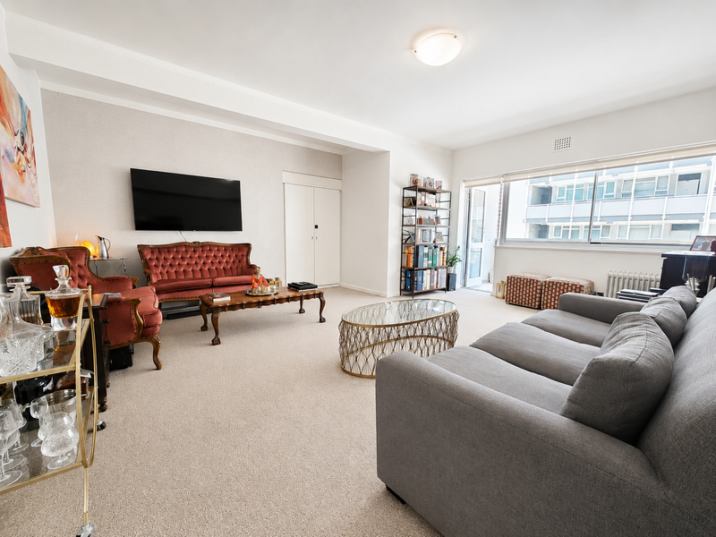 A spacious living room featuring neutral carpeting, a built-in storage cupboard, a radiator, and large windows providing natural light.