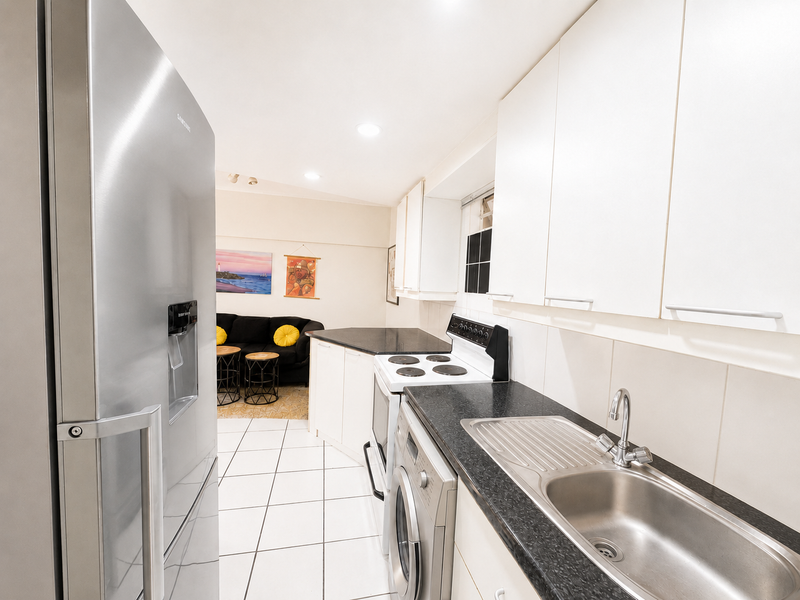 Open-plan kitchen featuring white cabinetry, dark countertops, stainless steel sink, and integrated appliances, flowing into a living area with tiled flooring throughout.