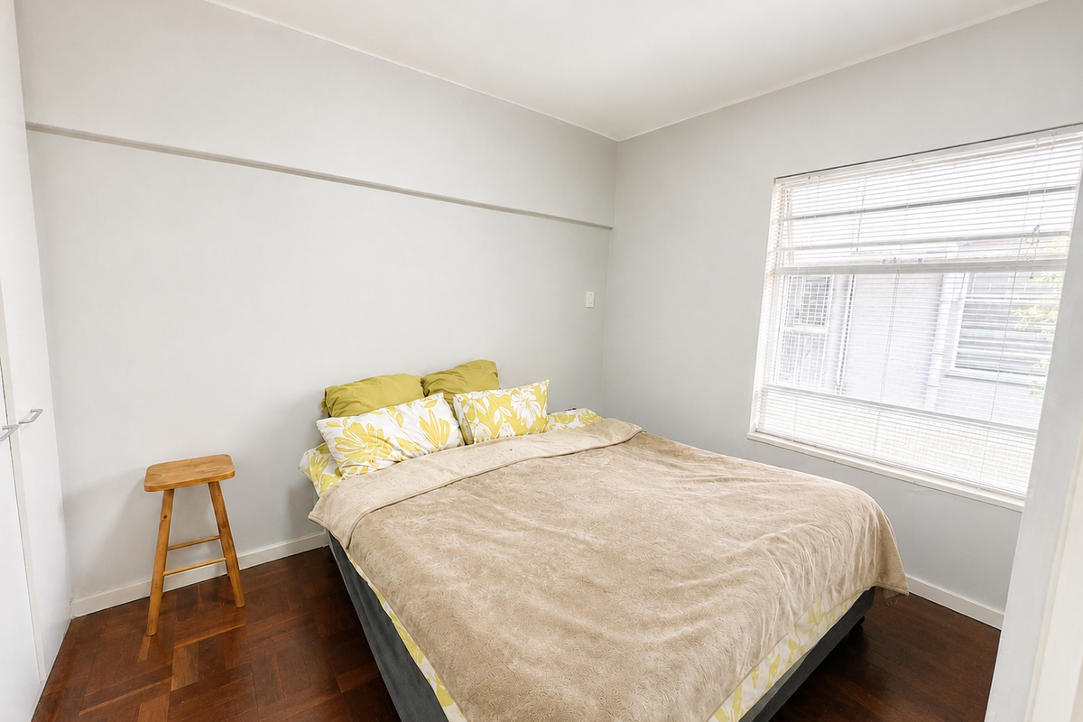 A bright bedroom featuring polished parquet flooring, neutral wall finishes, and a large window fitted with venetian blinds.