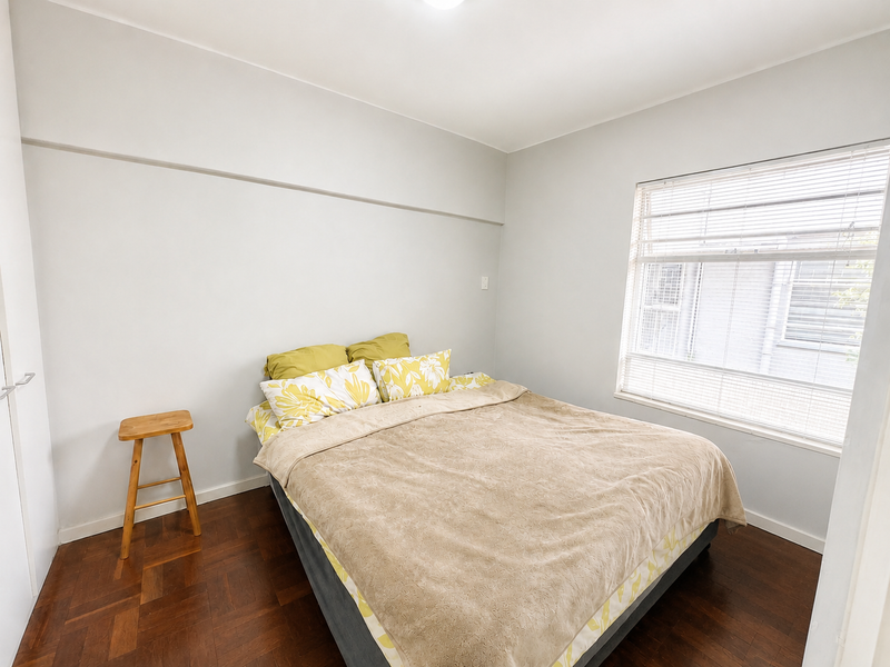 A bright bedroom featuring polished parquet flooring, neutral wall finishes, and a large window fitted with venetian blinds.