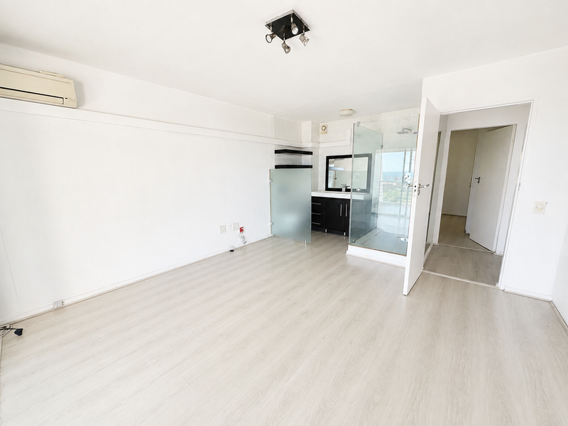 A bright, empty bedroom featuring laminate flooring, a wall-mounted air conditioner, and an open-plan en-suite bathroom with a glass shower enclosure.