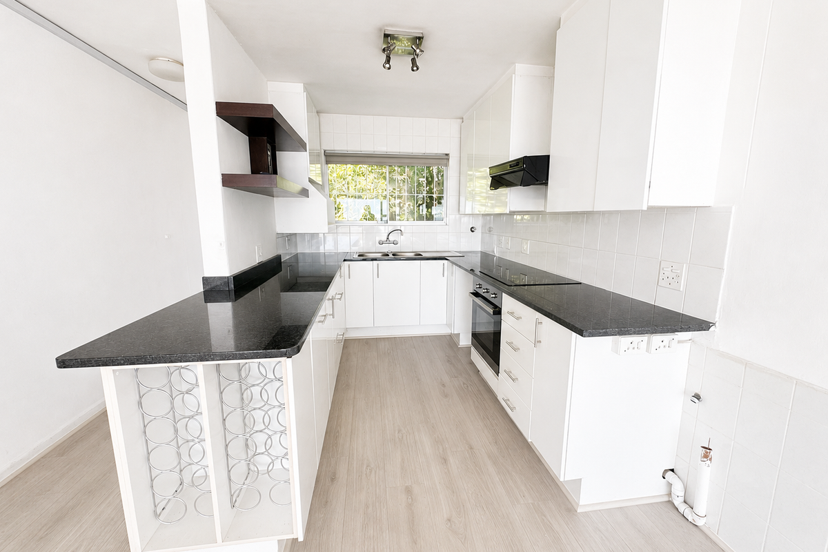 A clean, modern kitchen featuring white cabinetry, dark granite countertops, integrated appliances, and light wood-look laminate flooring in an open-plan layout.