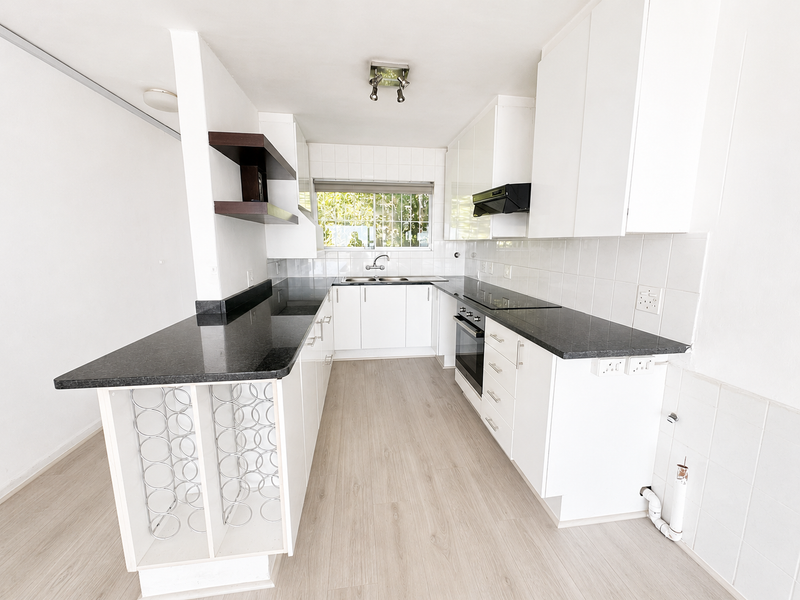 A clean, modern kitchen featuring white cabinetry, dark granite countertops, integrated appliances, and light wood-look laminate flooring in an open-plan layout.