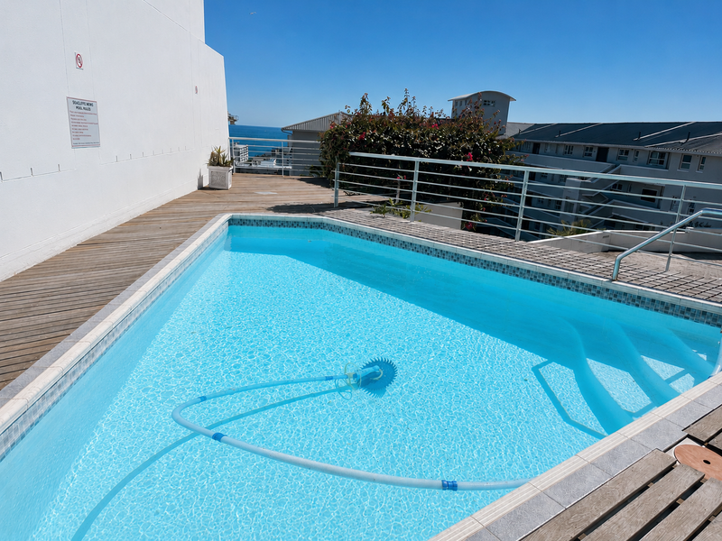 A well-maintained outdoor swimming pool area featuring wooden decking, mosaic tile detailing, and metal safety railings with views of the surrounding complex.
