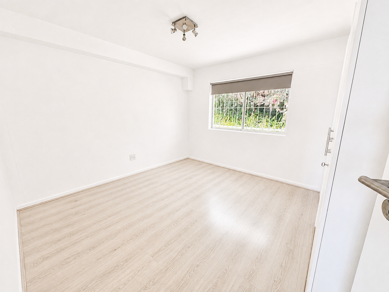 A bright, empty bedroom featuring light-toned laminate flooring, a neutral color palette, and a window fitted with security bars and a roller blind.