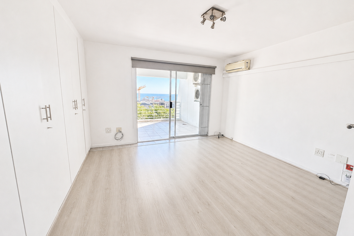 A bright, empty bedroom featuring light laminate flooring, built-in cupboards, an air conditioning unit, and a sliding door leading to a balcony with views.