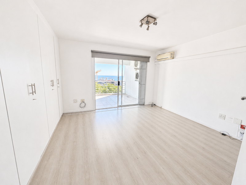 A bright, empty bedroom featuring light laminate flooring, built-in cupboards, an air conditioning unit, and a sliding door leading to a balcony with views.