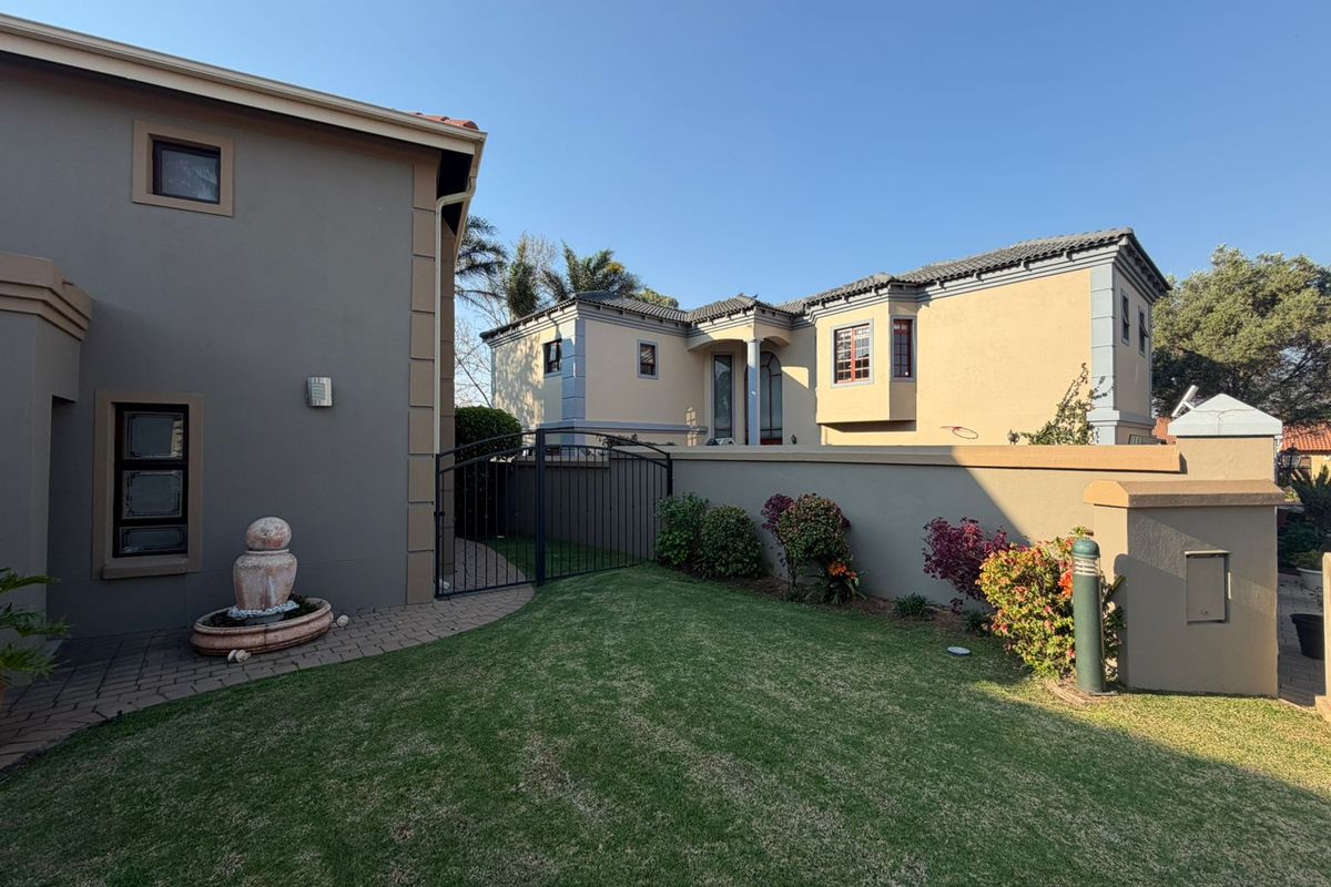A well-maintained rear garden area featuring a paved walkway, a stone fountain, a manicured lawn, and a secure wrought iron gate between two residential structures.