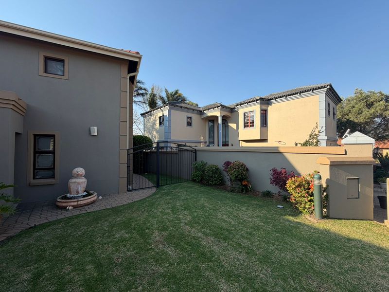 A well-maintained rear garden area featuring a paved walkway, a stone fountain, a manicured lawn, and a secure wrought iron gate between two residential structures.