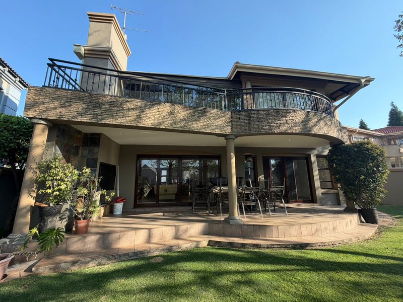 Spacious rear patio featuring stone cladding, tiled flooring, and a large upper-level balcony with wrought iron railings, overlooking the garden.