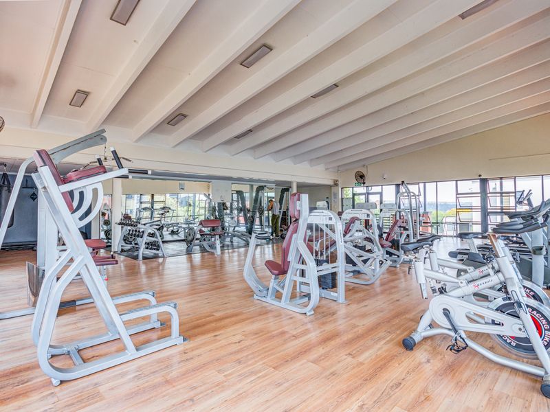 A spacious, well-lit gym facility featuring laminate flooring, mirrored walls, and exposed beam ceilings, equipped with various fixed strength and cardio training machines.