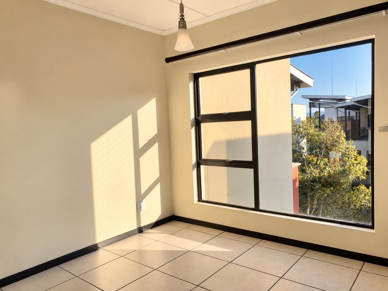 A bright, empty bedroom featuring neutral walls, tiled flooring, and a large window providing natural light and an exterior view.