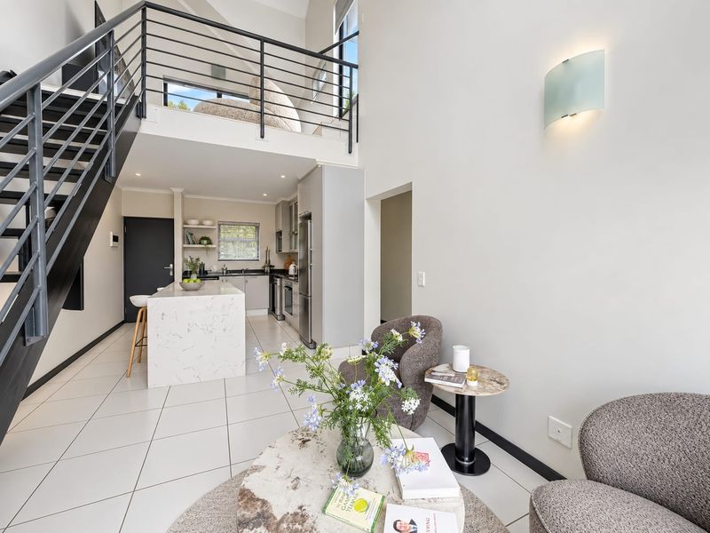 Modern open-plan living area featuring a sleek kitchen island, contemporary metal staircase, and a mezzanine level with high ceilings and tiled flooring.