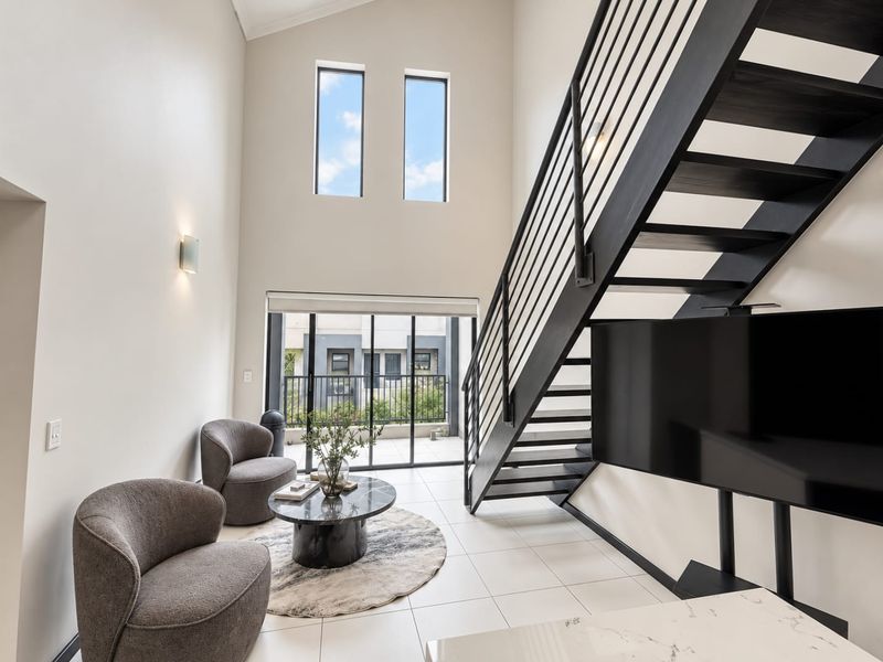 Modern living area featuring a striking black metal staircase, high vaulted ceilings, large windows, and tiled flooring with direct access to a balcony.