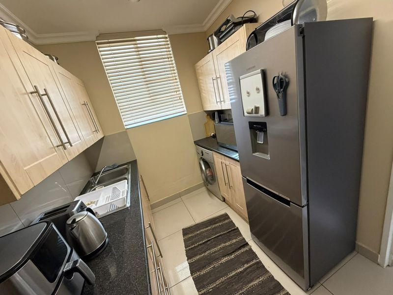 A functional galley-style kitchen featuring light wood cabinetry, dark countertops, tiled flooring, and integrated laundry plumbing.
