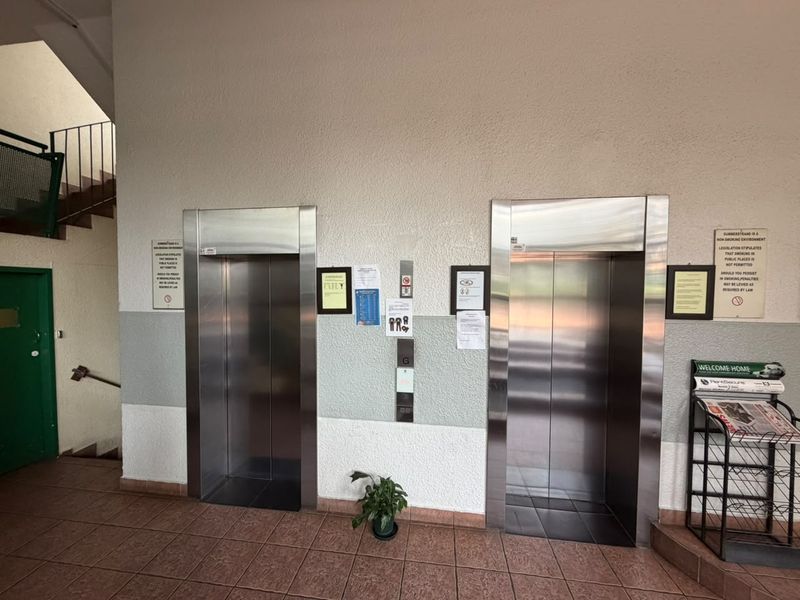 A communal apartment building hallway featuring dual stainless steel elevators, a staircase, and tiled flooring.