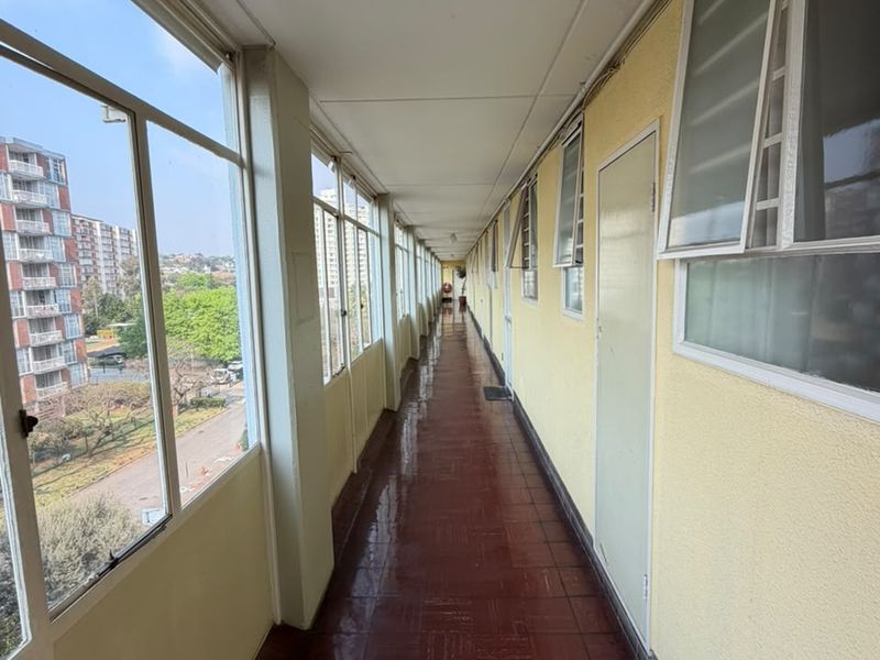 A long, sunlit apartment hallway featuring tiled flooring, large windows with exterior views, and multiple access doors along the corridor.