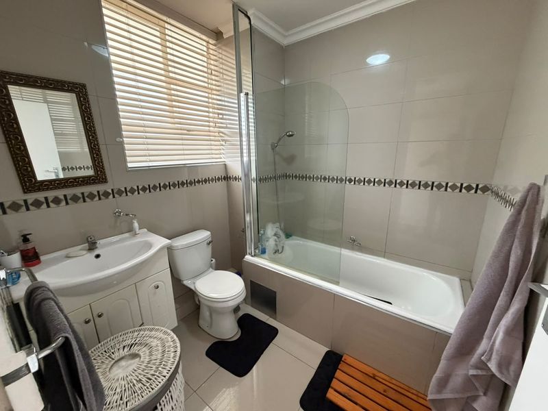A functional bathroom featuring a vanity cabinet, toilet, and a bathtub with a glass shower screen, finished with neutral wall and floor tiles.