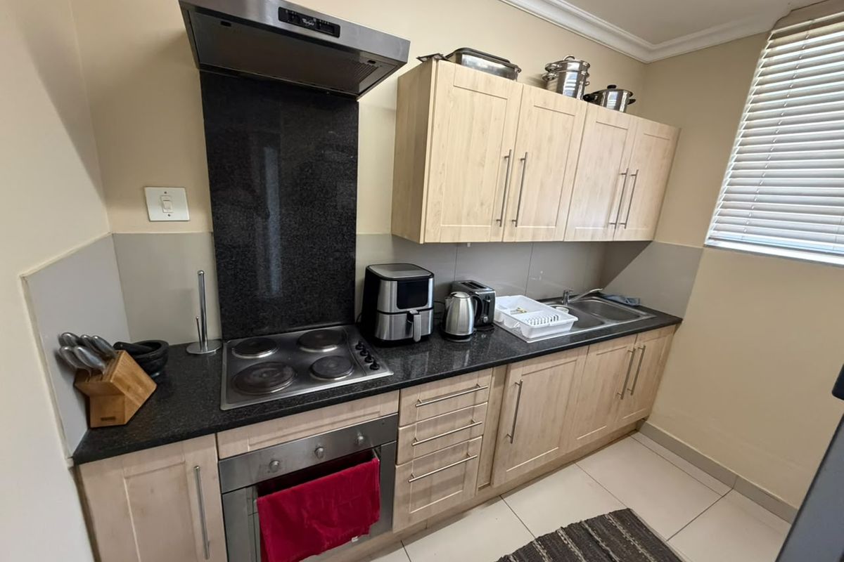 A functional kitchen featuring light wood cabinetry, granite countertops, a built-in electric hob, oven, and extractor fan, complemented by tiled flooring and window blinds.