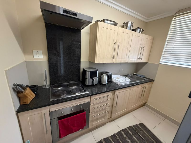 A functional kitchen featuring light wood cabinetry, granite countertops, a built-in electric hob, oven, and extractor fan, complemented by tiled flooring and window blinds.