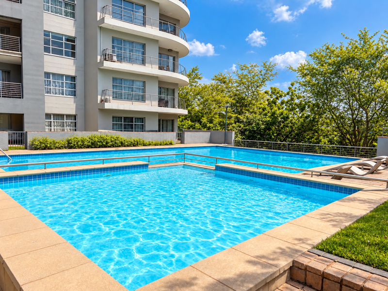 A well-maintained swimming pool area featuring tiled coping and metal safety railings, set against a modern multi-story apartment building with balconies.