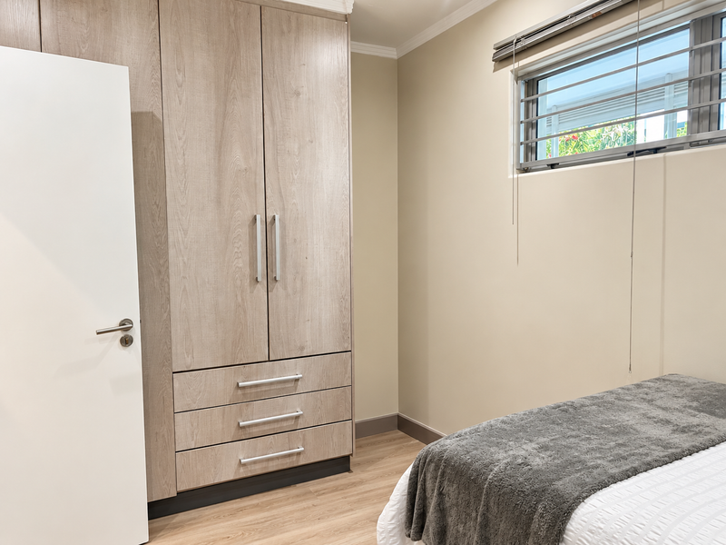 A well-maintained bedroom featuring light wood-grain built-in wardrobes, laminate flooring, and a window fitted with security bars and blinds.