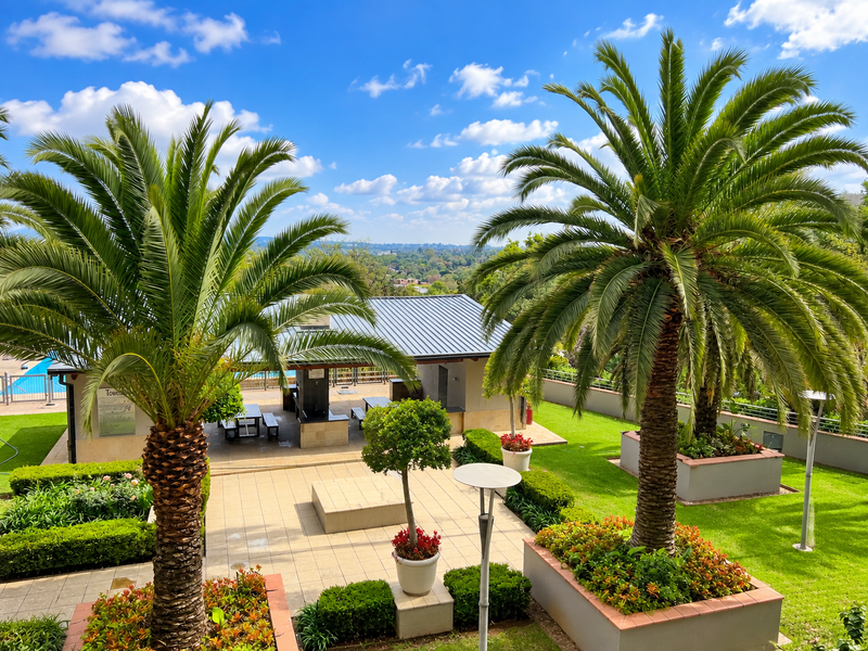 Well-maintained communal pool area featuring a covered patio with built-in braai facilities, surrounded by landscaped gardens and mature palm trees.