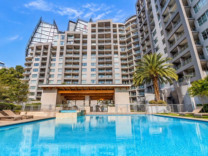 A well-maintained swimming pool area featuring a covered patio structure, metal fencing, and landscaped surroundings within a modern apartment complex.