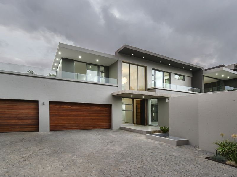 Modern double-story cluster home featuring a paved driveway, double wooden garage doors, and contemporary architectural design with glass balustrades.