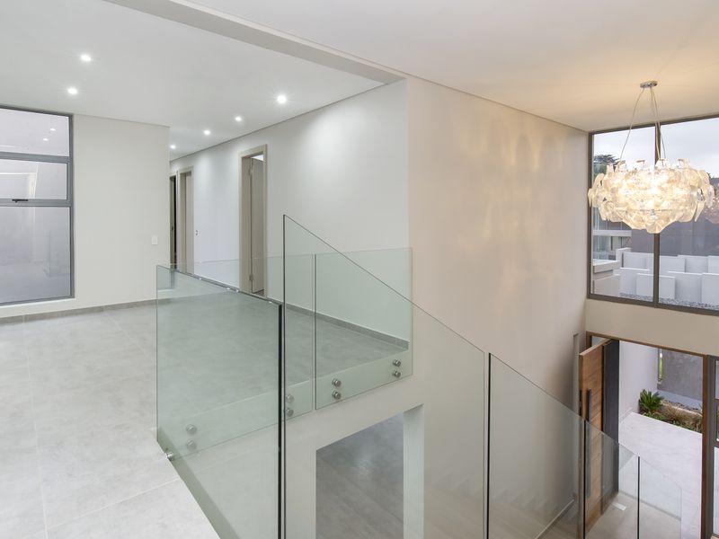 Modern upper-level landing area featuring a sleek glass balustrade, large format floor tiles, and recessed lighting overlooking the entrance foyer.