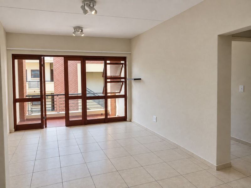 A bright, empty living area featuring neutral tiled floors and large timber-framed glass doors opening onto a balcony, providing ample natural light.