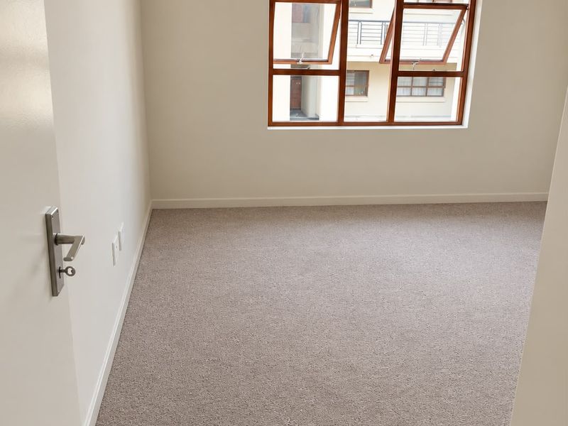A clean, empty bedroom featuring neutral carpeting, white walls, and a timber-framed window overlooking the complex exterior.