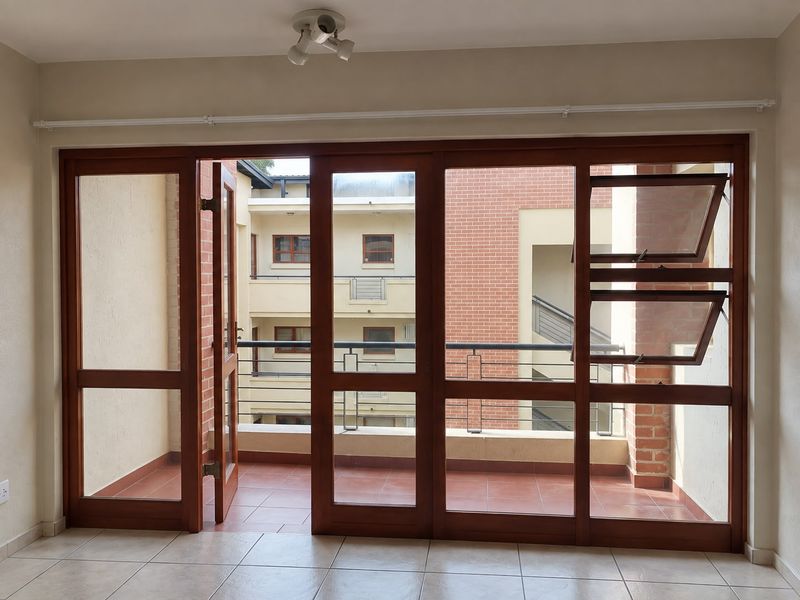 A covered balcony featuring timber-framed glass doors, tiled flooring, and a metal balustrade, overlooking the apartment complex courtyard.