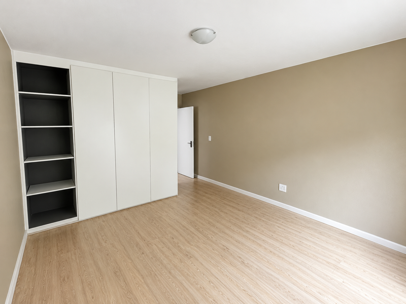 A bright, empty bedroom featuring modern laminate flooring, neutral wall tones, and a large built-in wardrobe with integrated shelving.