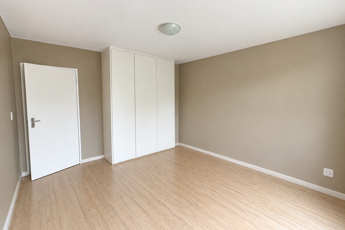 A bright, empty bedroom featuring light-toned laminate flooring, neutral wall paint, and a clean, white built-in wardrobe.
