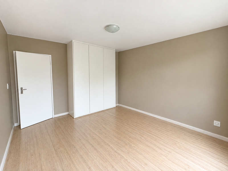 A bright, empty bedroom featuring light-toned laminate flooring, neutral wall paint, and a clean, white built-in wardrobe.