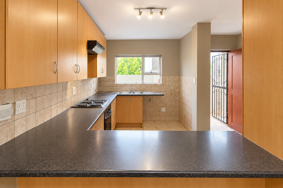 A functional kitchen featuring light wood-finish cabinetry, dark laminate countertops, built-in appliances, and tiled splashbacks, with direct access to an exterior area.