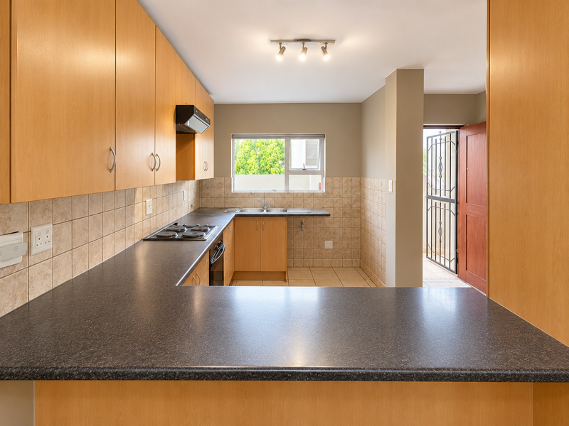 A functional kitchen featuring light wood-finish cabinetry, dark laminate countertops, built-in appliances, and tiled splashbacks, with direct access to an exterior area.