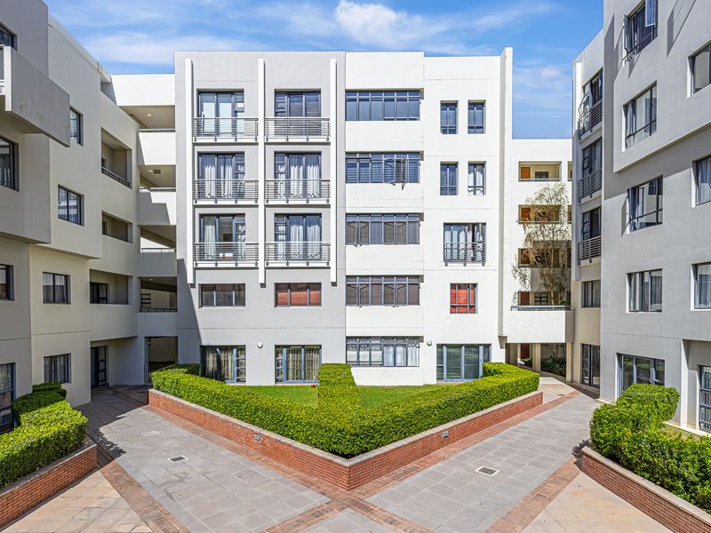 A well-maintained apartment complex courtyard featuring paved walkways, manicured hedges, and brick planter walls set against a modern multi-story residential building facade.