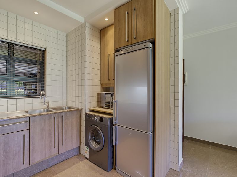 A well-maintained scullery featuring modern wood-finish cabinetry, a stainless steel double sink, tiled splashbacks, and dedicated space for laundry and refrigeration appliances.