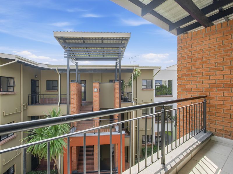 A well-maintained balcony featuring tiled flooring, a metal balustrade, and exposed brickwork, overlooking the apartment complex courtyard.