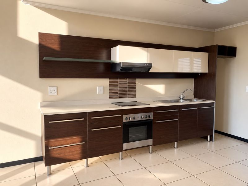 A modern, clean kitchen featuring dark wood-grain cabinetry, light countertops, integrated oven and hob, and neutral tiled flooring in an open-plan layout.