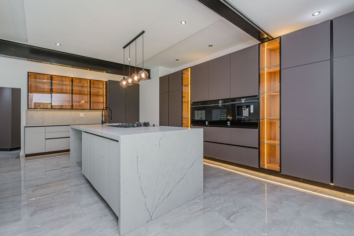 Modern luxury kitchen featuring a large stone island, integrated high-end appliances, custom cabinetry with ambient lighting, and polished marble-look floor tiles.