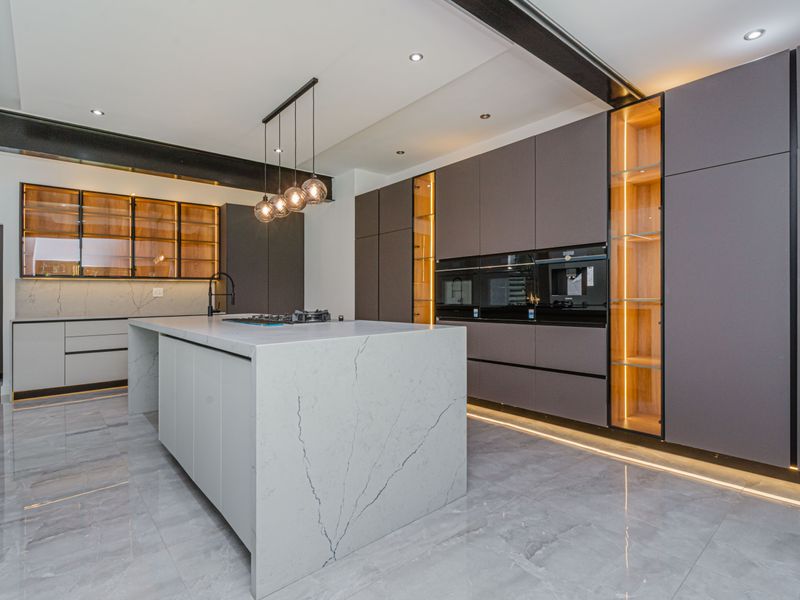 Modern luxury kitchen featuring a large stone island, integrated high-end appliances, custom cabinetry with ambient lighting, and polished marble-look floor tiles.