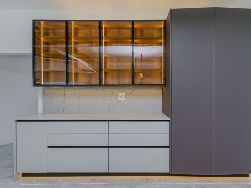 Modern kitchen featuring sleek handleless cabinetry, stone countertops with matching backsplash, and illuminated glass-front display shelving for a sophisticated, contemporary finish.