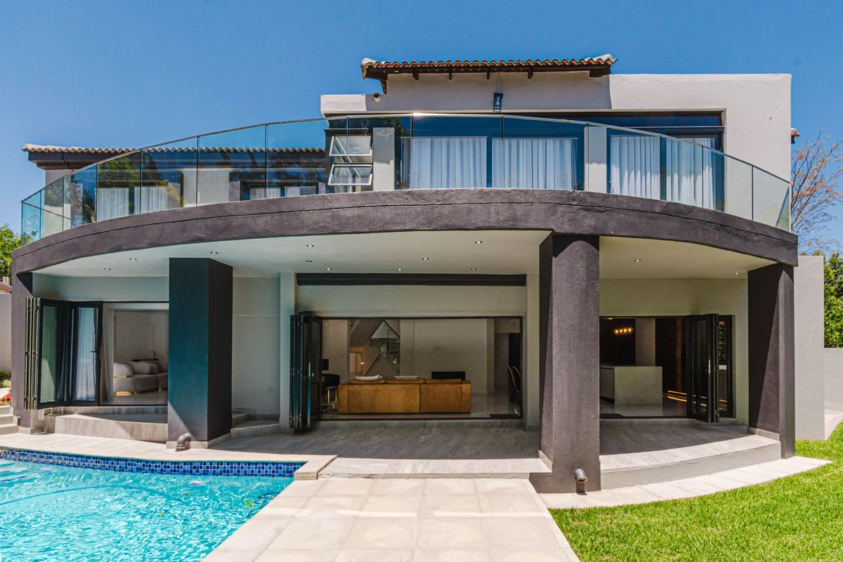 Rear elevation featuring a modern covered patio, glass balustrade balcony, and a swimming pool with landscaped garden access.