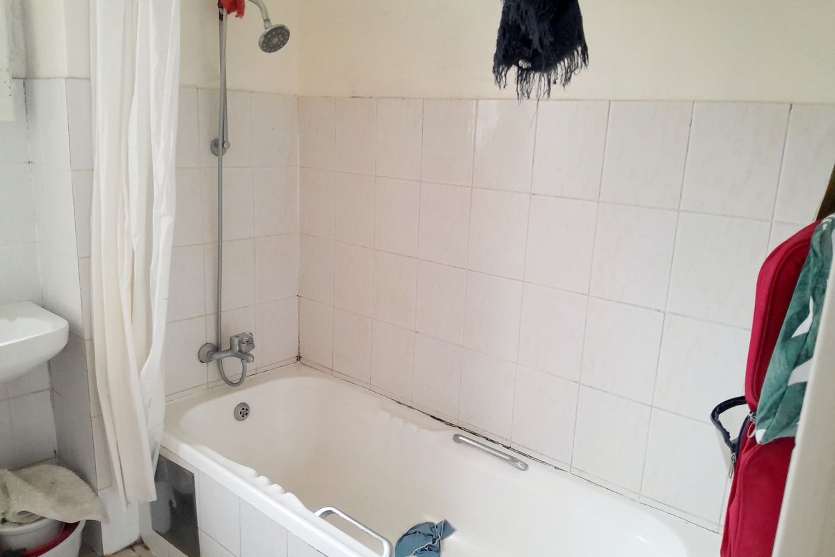 A functional bathroom featuring a bathtub with an integrated shower head, wall tiling, and a basin, currently showing signs of wear.