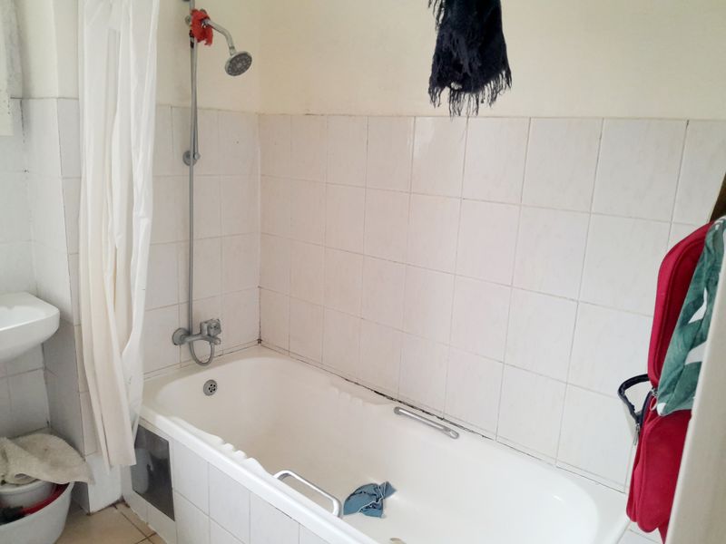 A functional bathroom featuring a bathtub with an integrated shower head, wall tiling, and a basin, currently showing signs of wear.