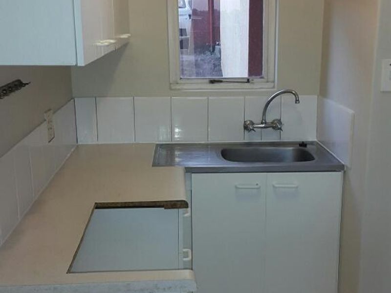 A compact kitchen area featuring white built-in cabinetry, a stainless steel sink, tiled splashback, and ceramic floor tiles, currently awaiting appliance installation.