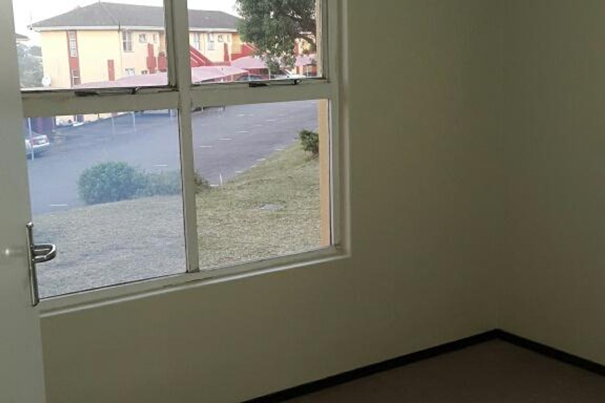 A vacant bedroom featuring neutral walls, carpeted flooring, and a large window providing natural light and views of the complex grounds.
